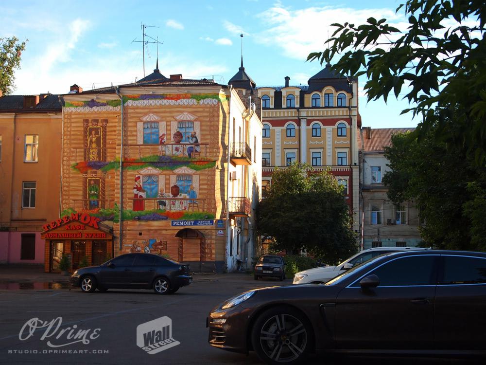Budynek z muralem imitującym drewniany dom z postaciami, napisami w języku rosyjskim i zaparkowanymi samochodami na pierwszym planie w słoneczny dzień.