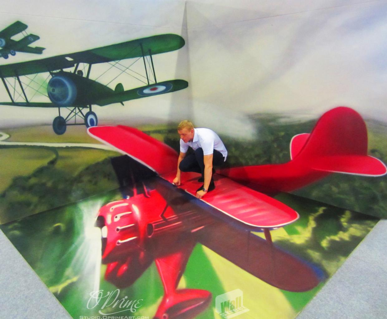Mężczyzna w kucki na iluzji 3D czerwonego samolotu, z zielonymi samolotami w tle. Perspektywa z góry, jasne oświetlenie, widoczne logo studia.