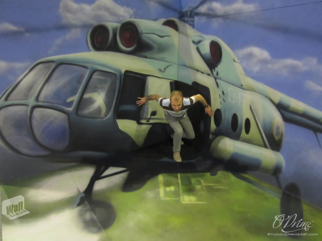 Mężczyzna pozuje na tle iluzji optycznej helikoptera, sprawiając wrażenie wyskakującego z niego. Nietypowe ujęcie, mural artystyczny.