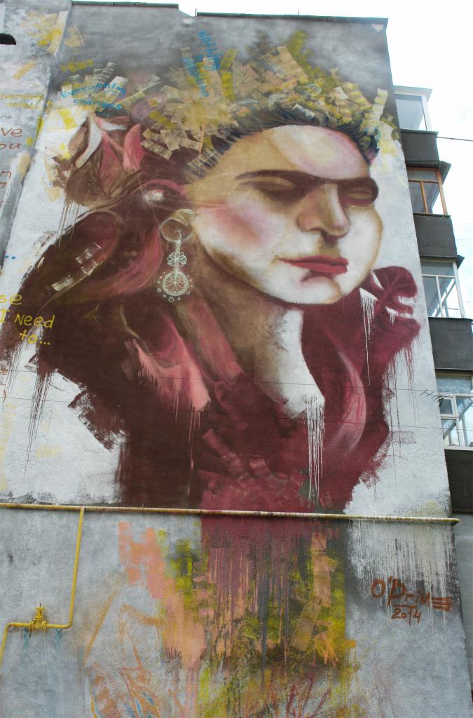 Mural na ścianie budynku przedstawia portret kobiety z kolczykiem i nakryciem głowy z fragmentów dokumentów, w brązowo-czerwonej kolorystyce.