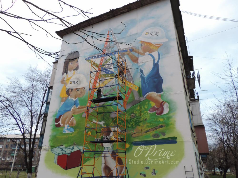 Mural na ścianie bloku przedstawiający dzieci budujące konstrukcję z metalowych elementów, malowany z użyciem rusztowania. Sygnowany 'O'Prime Studio.OprimeArt.com'.