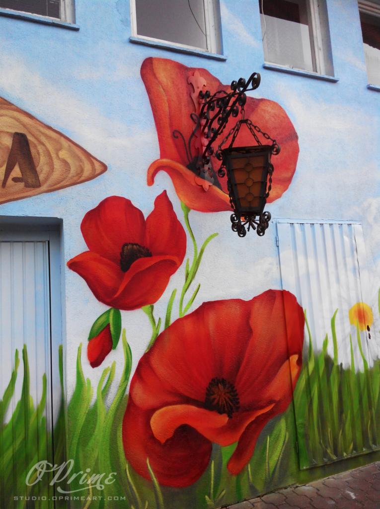 Malowidło ścienne z motywem czerwonych maków, drewniany szyld i ozdobna latarnia na tle błękitnego nieba. Artystyczna elewacja budynku.