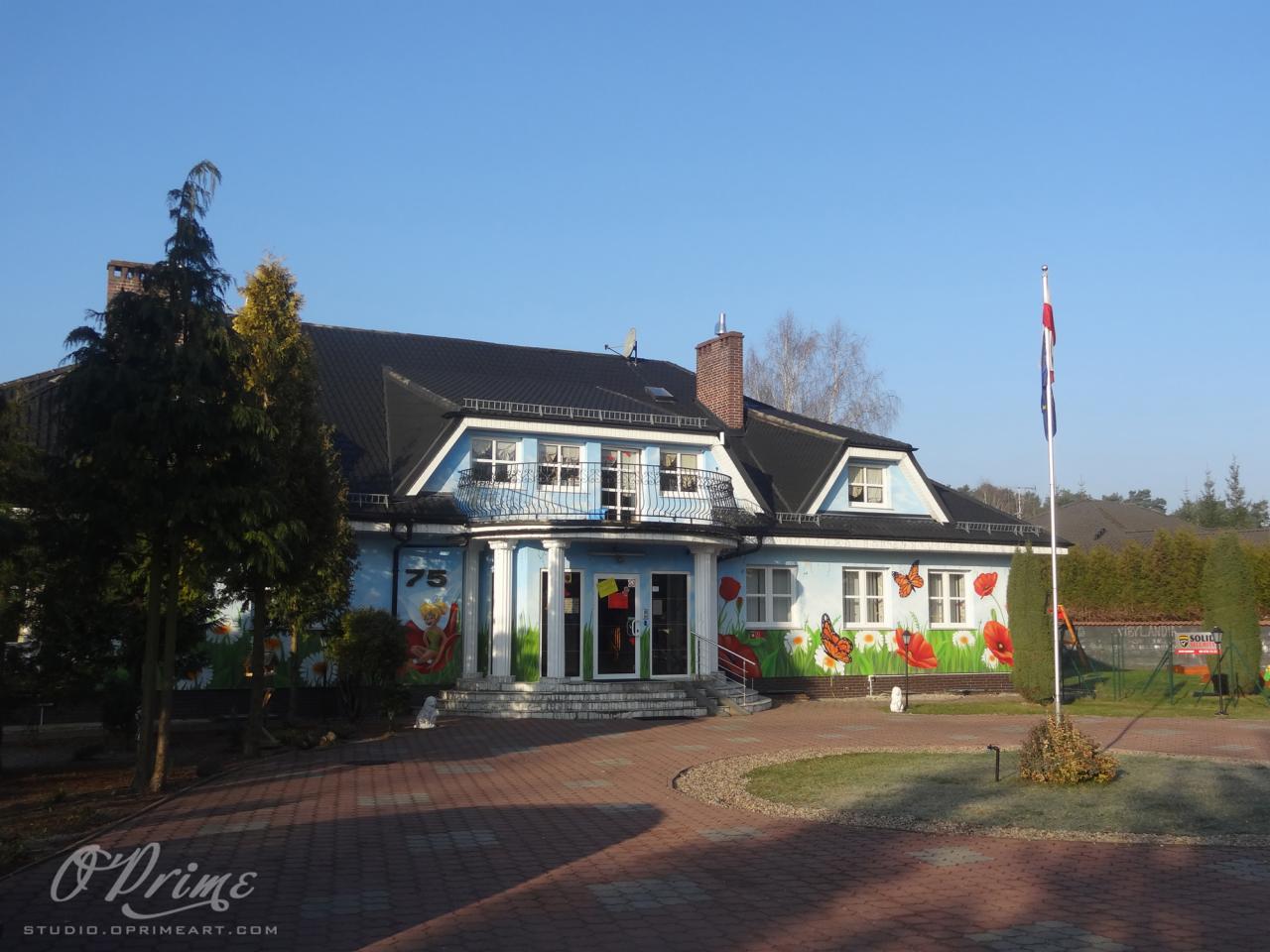 Budynek z numerem 75 i ręcznie malowanymi kwiatami na elewacji. Dwuspadowy dach, balkon, flaga. Kostka brukowa na podjeździe. Słoneczny dzień.