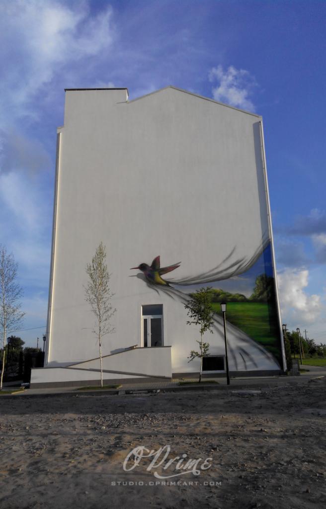 Malowidło ścienne z kolibrem i krajobrazem na białym budynku, widoczne drzewa i niebo. Ujęcie z dołu podkreśla monumentalność dzieła.