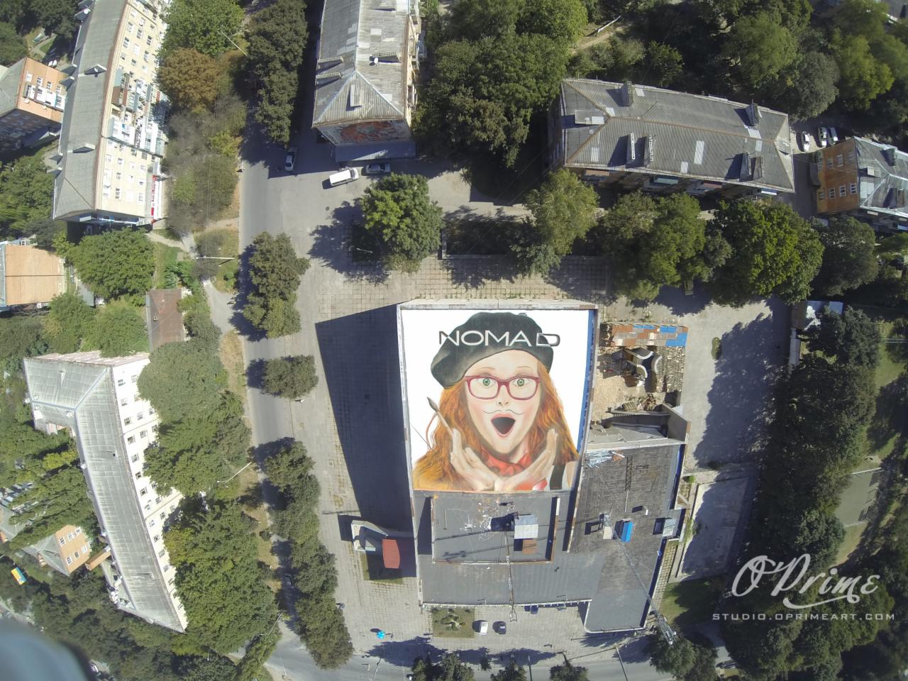 Widok z góry na budynek z muralem przedstawiającym kobietę w okularach i beretce, obok zieleń miejska i zabudowa. Ujęcie z lotu ptaka.