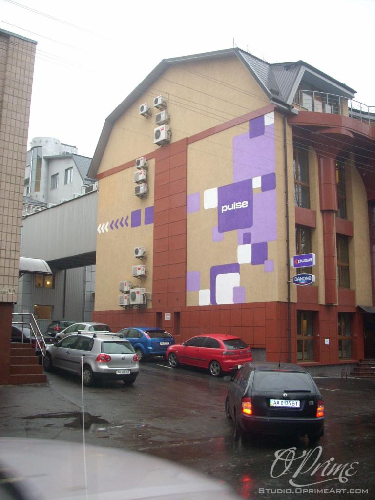 Budynek z logo 'pulse' i 'DANONE' w fioletowo-białej kolorystyce, na tle zaparkowanych samochodów w deszczowy dzień. Widoczne klimatyzatory na elewacji.