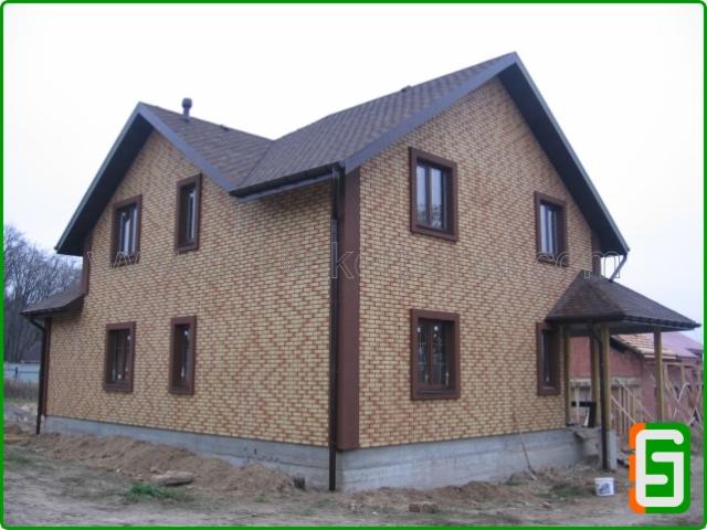 Dwukondygnacyjny dom z elewacją z cegły klinkierowej w odcieniach beżu i brązu, z brązowymi ramami okiennymi i ciemnobrązowym dachem. Widok z zewnątrz.