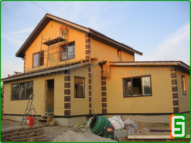 Budowa domu jednorodzinnego z cegły klinkierowej, widoczne rusztowanie, worki z zaprawą i betoniarka. Dom z brązowymi obramowaniami okien i dachu.