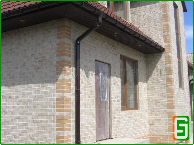 Elewacja budynku wykończona jasną cegłą klinkierową z brązowymi akcentami, widoczne okno i drzwi zabezpieczone folią, brązowa rynna i dachówka.