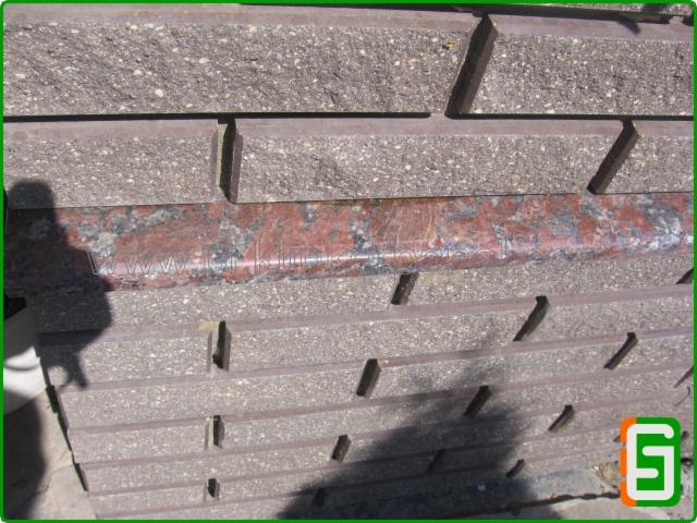 Ułożone obok siebie, szare i czerwone elementy kamienne z rowkami, prawdopodobnie krawężniki lub obrzeża chodnikowe. Widoczny cień osoby.