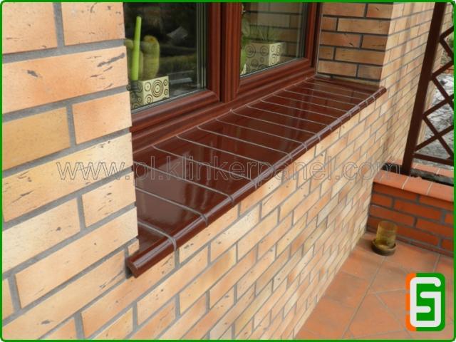 Klinkierowa obudowa okna z brązowym parapetem i ceglaną ścianą. Widoczny fragment balustrady i doniczka na parapecie.