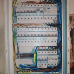 Otwarta skrzynka elektryczna z zamontowanymi bezpiecznikami i podłączonymi przewodami w różnych kolorach, umieszczona na ścianie wewnątrz budynku.