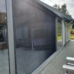 Zewnętrzna, plisowana siatka przeciw owadom zamontowana na tarasie domu jednorodzinnego, widoczna z zewnątrz, z fragmentem drabiny aluminiowej w dolnym rogu.