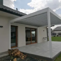 Nowoczesna, biała pergola lamelowa przylegająca do domu z dużymi oknami przesuwnymi, na szarym tarasie z ułożonym drewnem opałowym.