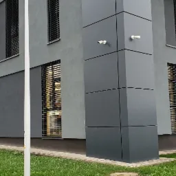 Szary budynek biurowy z zamontowanymi poziomymi osłonami okiennymi, widoczne kamery monitoringu na rogu budynku.