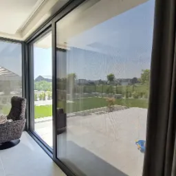 Nowoczesne przesuwne okno tarasowe z szarą, transparentną przesłoną przeciwsłoneczną, widok z wnętrza na taras i ogród, fotel obok okna.