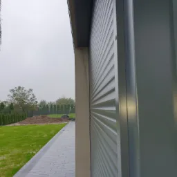 Szare, zewnętrzne osłony okienne zamknięte, widok z boku, na tle trawnika i elementów krajobrazu w pochmurny dzień.