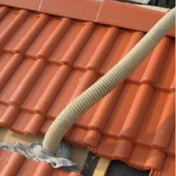 Wąż do wdmuchiwania materiału izolacyjnego wystający z otworu w czerwonej dachówce, zabezpieczony szarą tkaniną.