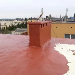 Czerwony dach pokryty hydroizolacyjną powłoką z widocznym kominem i anteną satelitarną na tle zabudowy miejskiej w pochmurny dzień.