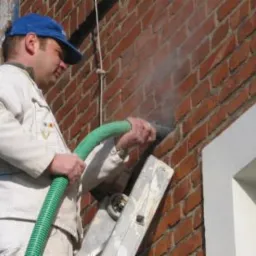 Pracownik w niebieskiej czapce aplikuje materiał izolacyjny granulatem w ścianę z czerwonej cegły za pomocą zielonego węża, stojąc na drabinie.