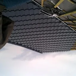 Układanie grafitowej dachówki ceramicznej na drewnianej więźbie dachowej, widok z dołu na fragment dachu i robotnika w brązowej bluzie.