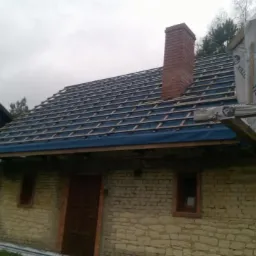 Dom w trakcie montażu łat i kontrłat na niebieskiej membranie dachowej, widoczny komin z cegły, obok budynek gospodarczy z eternitem, pochmurne niebo.