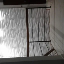 Ściana z dekoracyjnymi panelami 3D, częściowo zasłonięta przez konstrukcję schodów, oświetlona z lewej strony, z widocznymi płytkami na sąsiedniej ścianie.