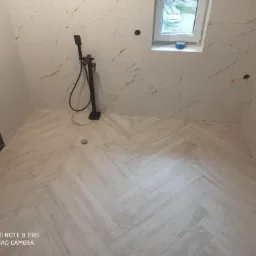 Wykończona łazienka z jasnymi płytkami imitującymi marmur z delikatnymi złotymi żyłami na ścianach i podłodze ułożonej w jodełkę, czarna armatura natryskowa, okno z nieukończonym wykończeniem.