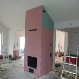 Konstrukcja kominka obudowana płytami kartonowo-gipsowymi w trakcie montażu, widoczne otwory wentylacyjne i wkład kominkowy, surowy beton na podłodze, drabiny i narzędzia w tle.