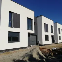 Nowoczesne, szeregowe domy jednorodzinne z jasną elewacją i szarymi akcentami, widoczne w stanie surowym, z piaskiem i materiałami budowlanymi na pierwszym planie, błękitne niebo w tle.