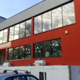 Czerwony budynek z dużymi oknami, częściowo wykończony, widoczne materiały budowlane, reklama 'meble bodzio' nad wejściem.