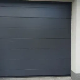Zamknięta, segmentowa brama garażowa w kolorze antracytowym, z poziomymi przetłoczeniami, zamontowana w otynkowanej wnęce budynku z szarym cokołem.