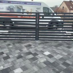 Szara, pozioma brama wjazdowa na posesję, z widocznym fragmentem firmowego samochodu z reklamą kamer i automatyki bram w tle.