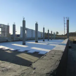 Budowa budynku żelbetowego: widok na fundamenty z ułożonym styropianem, słupy z wystającym zbrojeniem i fragment ściany z pustaków. W tle błękitne niebo i sprzęt budowlany.