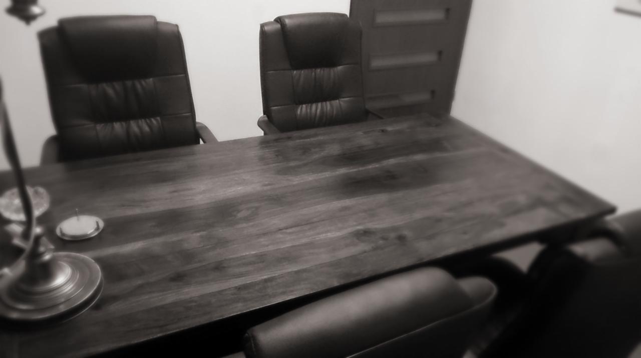 Czarno-białe zdjęcie pustego, ciemnego, drewnianego biurka z dwoma skórzanymi fotelami biurowymi i lampą biurkową w tle.