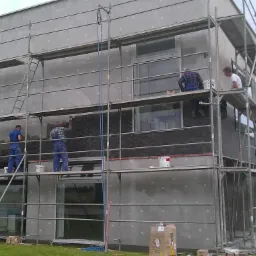 Prace elewacyjne na budynku: montaż izolacji na rusztowaniu. Czterech pracowników w kombinezonach. Widoczne materiały budowlane i narzędzia, teren zielony.