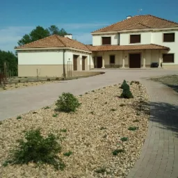 Elegancki dom z beżową elewacją i brązowymi oknami. Widoczny podjazd z kostki, kamienne rabaty i zadbana zieleń. Całość w słoneczny dzień.