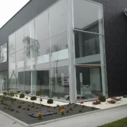 Nowoczesny budynek z dużymi przeszkleniami, widoczny pracownik malujący elewację, otoczenie z elementami architektury krajobrazu i chodnikiem.