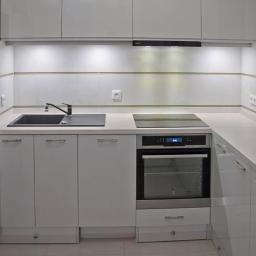 Lynx- Ryszard Suchora - Biała, nowoczesna kuchnia z czarnym zlewem i płytą indukcyjną w Poznaniu. Górne szafki podświetlone, minimalistyczne uchwyty, jasne płytki na ścianie.