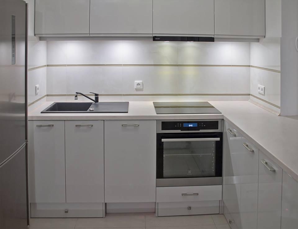 Biała, nowoczesna kuchnia z czarnym zlewem i płytą indukcyjną w Poznaniu. Górne szafki podświetlone, minimalistyczne uchwyty, jasne płytki na ścianie.