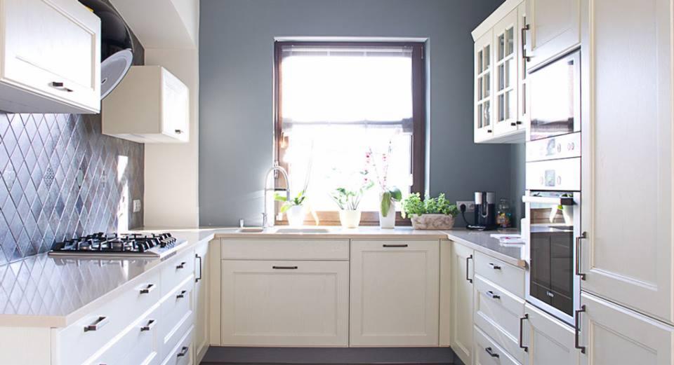 Jasna kuchnia w stylu klasycznym z szarymi płytkami na ścianie, widok na okno z roślinami. Ustawne szafki i szuflady z ciemnymi, podłużnymi uchwytami.