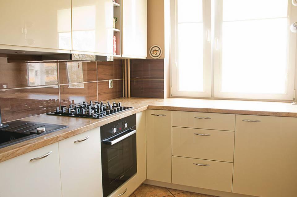Jasna kuchnia z beżowymi frontami, brązowym blatem i płytkami. Widoczna płyta grzewcza i piekarnik. Nad oknem dekoracyjny talerz.