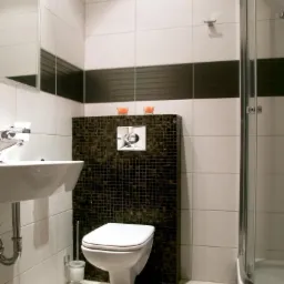 Nowoczesna łazienka z wiszącą toaletą na tle mozaiki, białą umywalką i kabiną prysznicową, akcentowana czarnym pasem płytek i świecami.