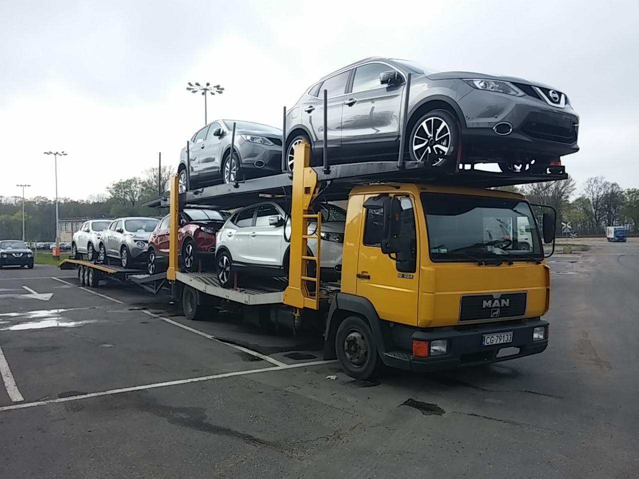 Żółta laweta MAN z naczepą, przewożąca pięć samochodów Nissan Qashqai w różnych kolorach, zaparkowana na parkingu. Widoczne drzewa i sygnalizacja kolejowa w tle.