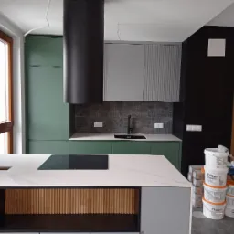 Wnętrze kuchni w trakcie wykańczania: zielone i szare szafki, czarny okap cylindryczny, wyspa z blatem imitującym marmur i drewnianymi lamelami, widoczne puszki farb Flugger i narzędzia malarskie.