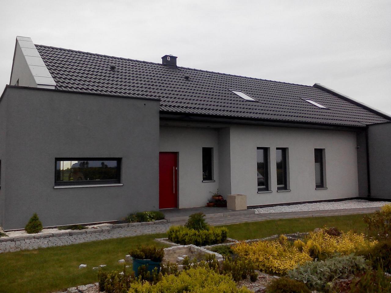 Nowoczesny dom jednorodzinny z grafitową dachówką i czerwoną stolarką drzwiową. Elewacja w odcieniach szarości, z zaakcentowanym oknem. Uwagę zwraca zadbany ogród.