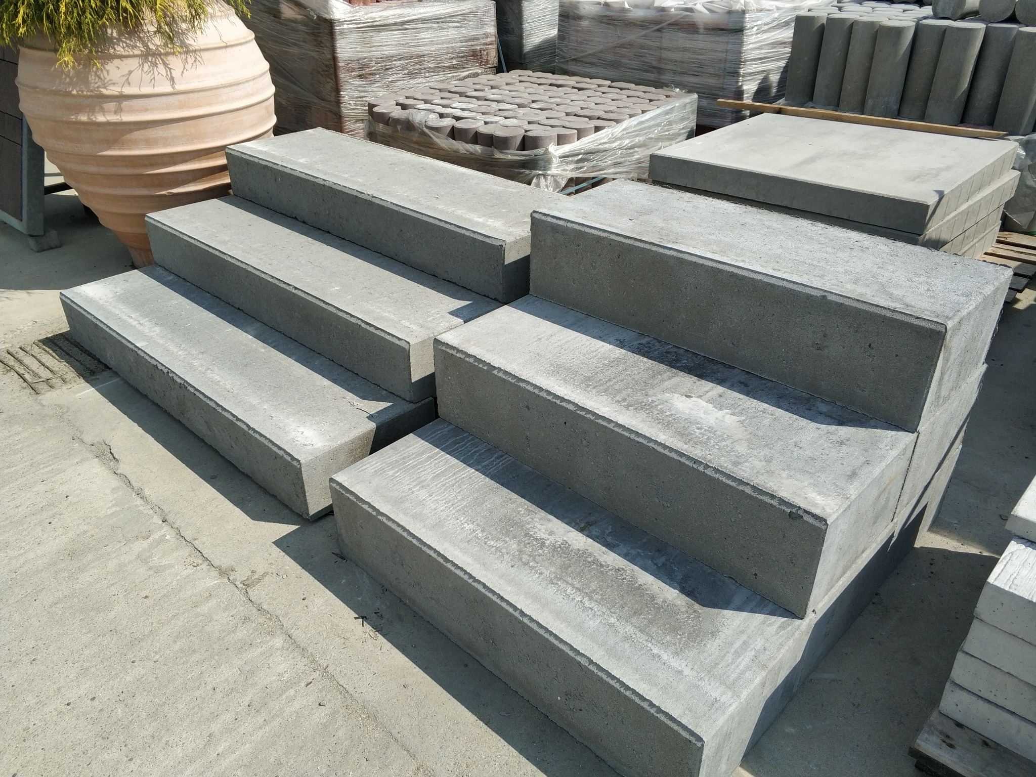 Stopnie schodowe ułożone z prefabrykowanych elementów betonowych, w tle donica z roślinami i zapakowane kostki brukowe.