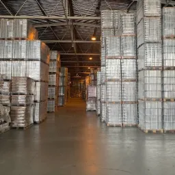 Wnętrze magazynu z rzędami palet z metalowymi pojemnikami, oświetlone lampami sufitowymi, widok w głąb korytarza.