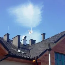 Dom z ciemną dachówką, kominami i anteną telewizyjną oraz zainstalowaną instalacją odgromową w postaci pionowych prętów, na tle błękitnego nieba z chmurami.
