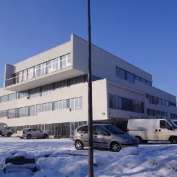 Nowoczesny, trzypiętrowy budynek biurowy z białej cegły, z dużymi oknami, widziany zimą. Na pierwszym planie ośnieżony parking z zaparkowanymi samochodami.